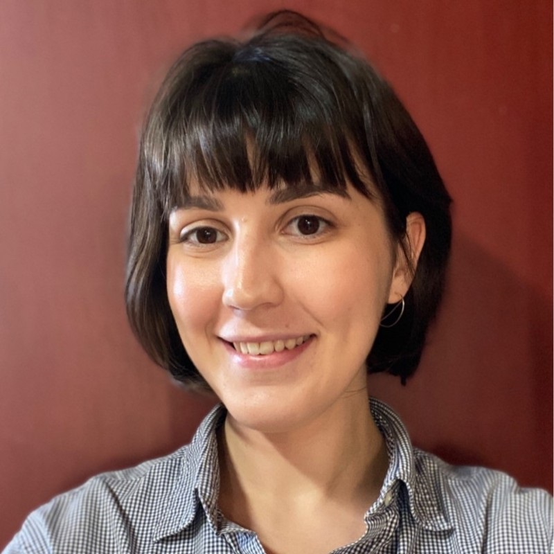 woman with short hair cut and bangs. She is wearing a blue-grey blouse and a small silver necklace