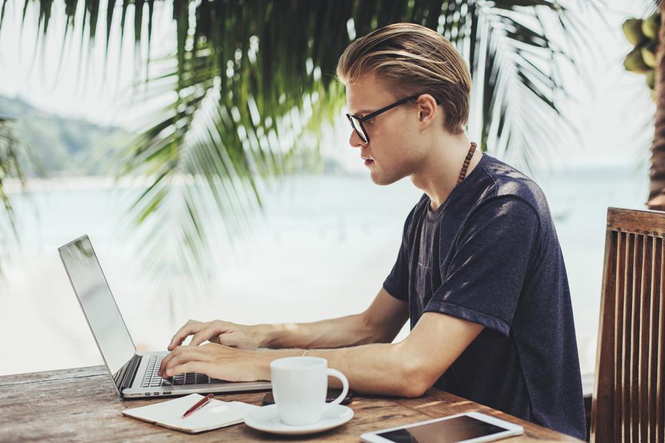 Man sits at a table, wearing a short sleeve shirt. He is working on his laptop and their is a blurbed background of palm trees and a beach
