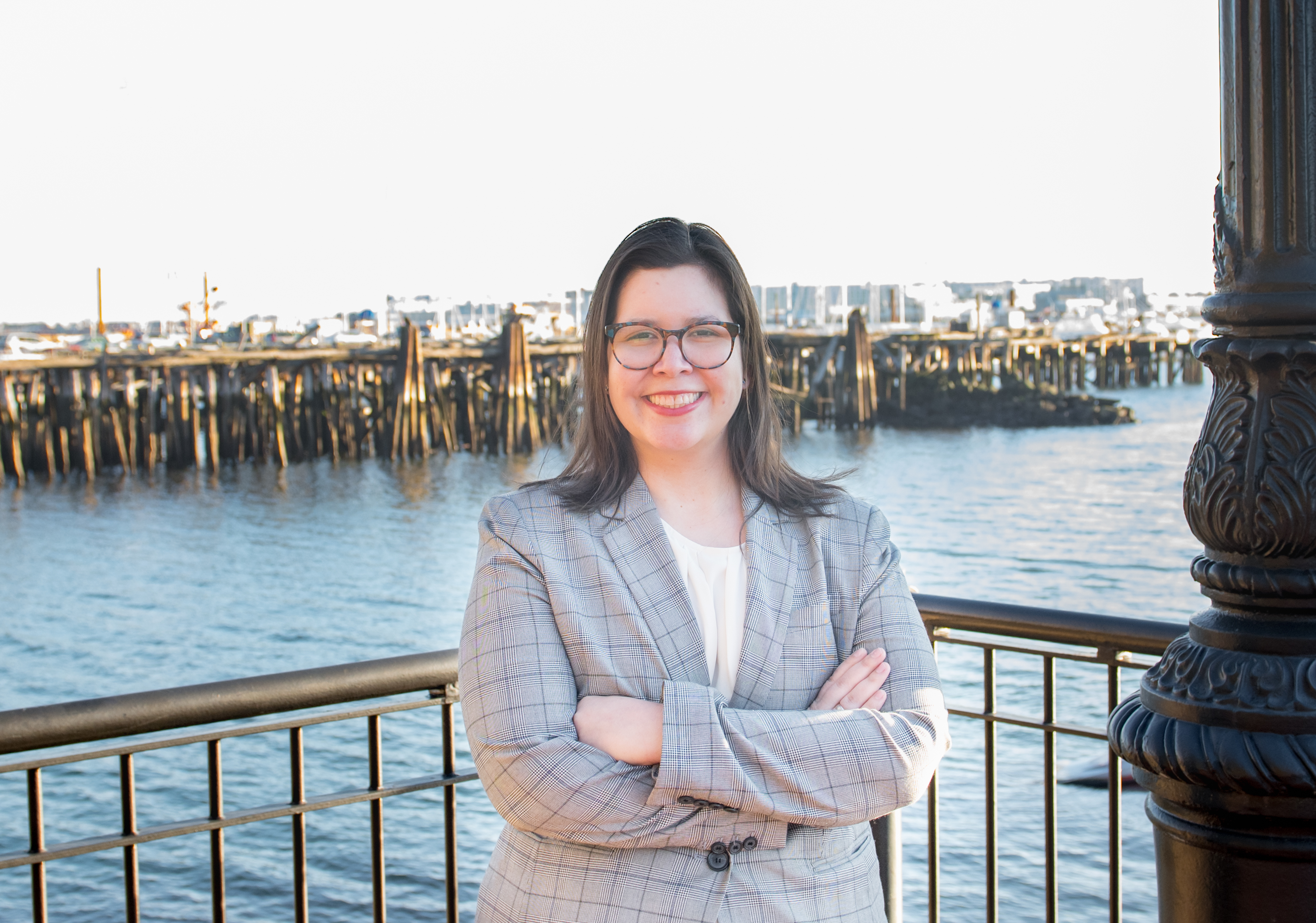 photo of woman standing in front of boston harbor. She is wearing a gray suit jacket and eye glasses. She is smiling. 